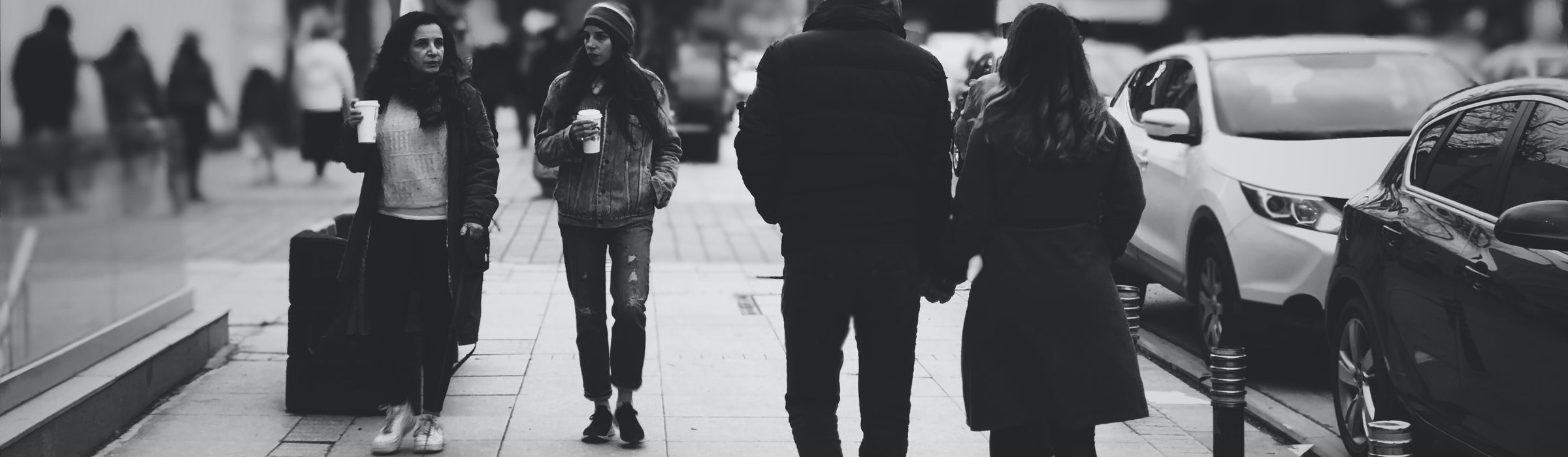 People walking along a busy city pavement, two of them holding coffee cups, in black and white