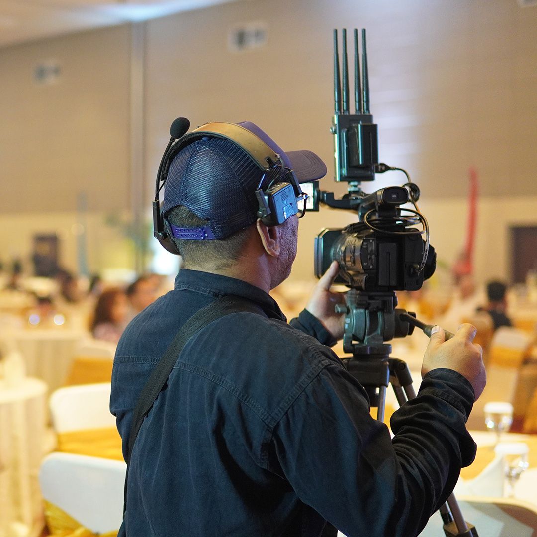 A camera operator in headphones filming a conference from a tripod in a busy function room