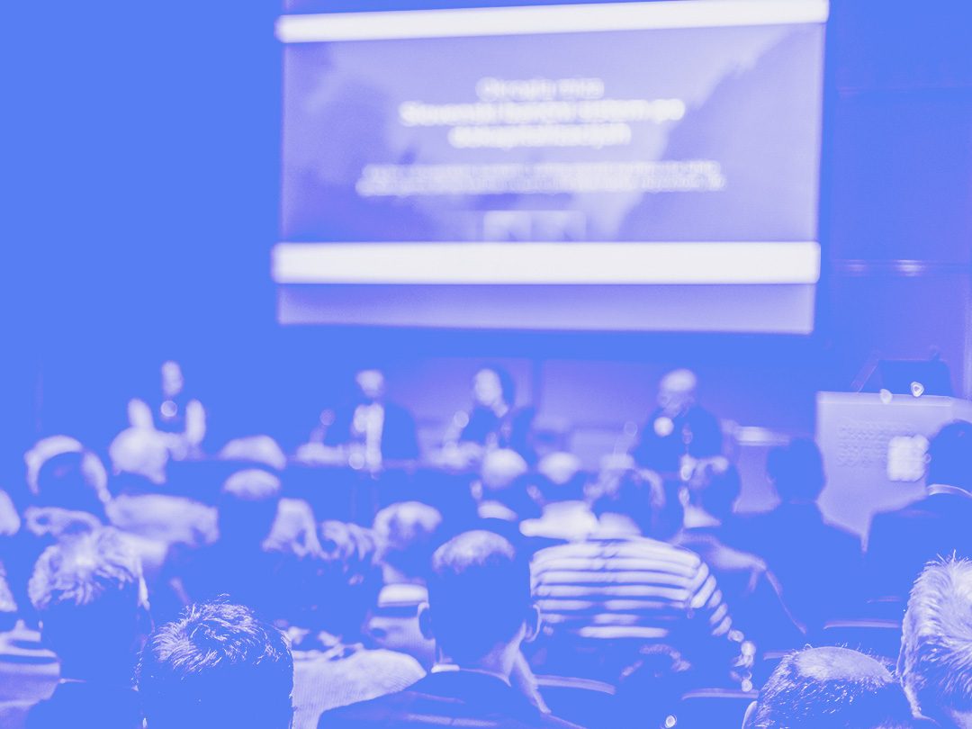 An audience watching a presenter on stage at a conference, tinted blue